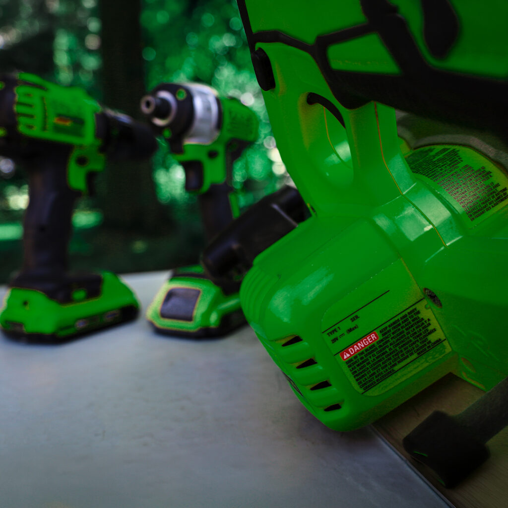 Green power saw with warning label in sharp focus outdoors, with a drill and another tool blurred in the background on white surface.