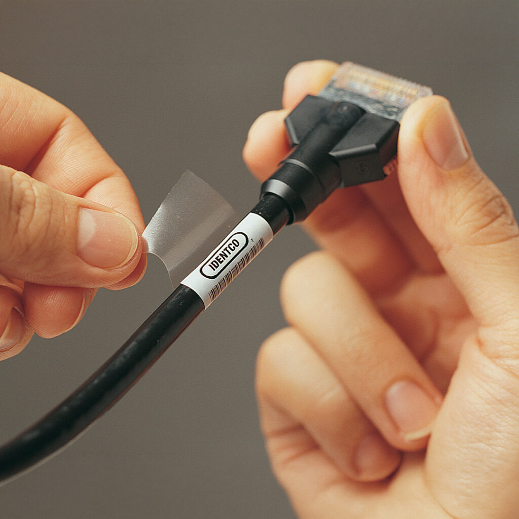 Hands placing a clear adhesive label over a white ID label marked IDENTO on a black cable attached to a rectangular connector.