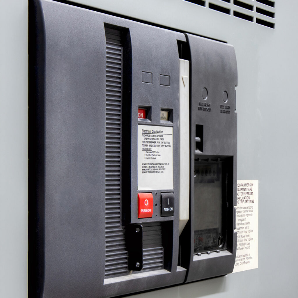 Close-up of gray electrical circuit breaker panel with switches, red ON button, white instruction label, and safety warnings on flat surface.