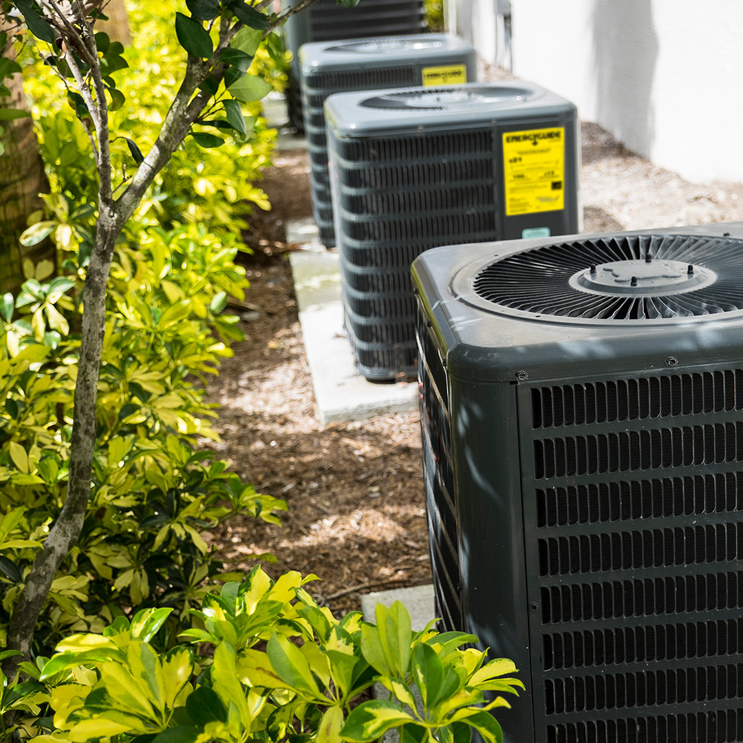 Outdoor air conditioning units on concrete pads next to a white building, bordered by green shrubs with sunlight filtering through leaves.