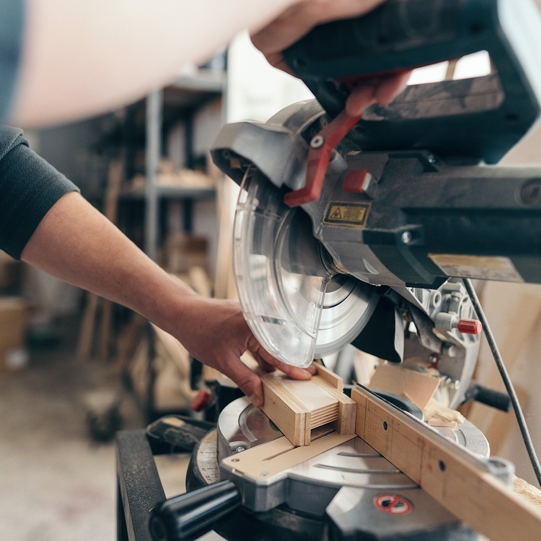 Person cutting wood with a circular miter saw in a workshop, shelves and woodworking tools visible in the background.