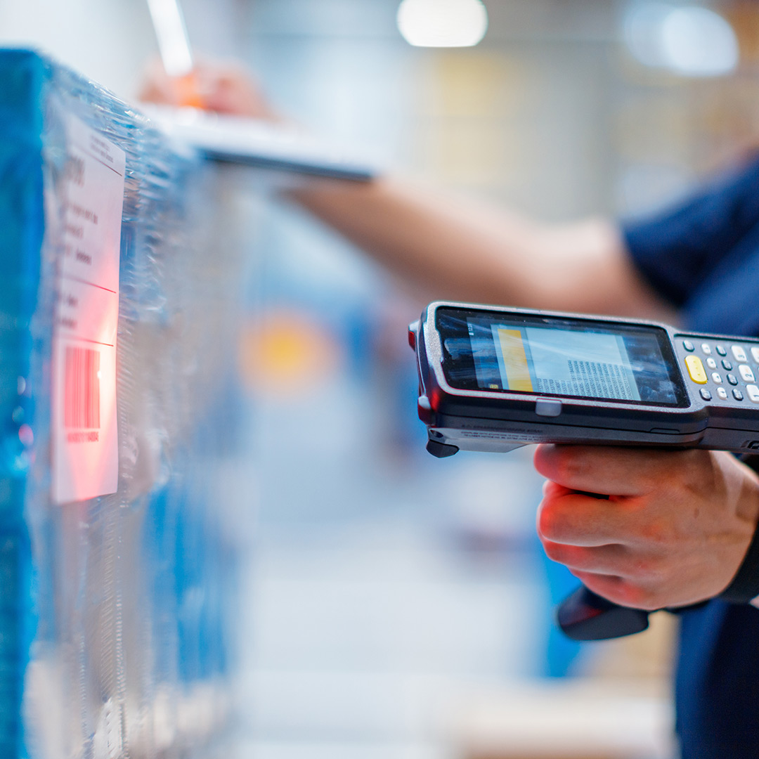 Warehouse worker scans barcode on a wrapped package with a handheld scanner while holding a clipboard and taking notes.