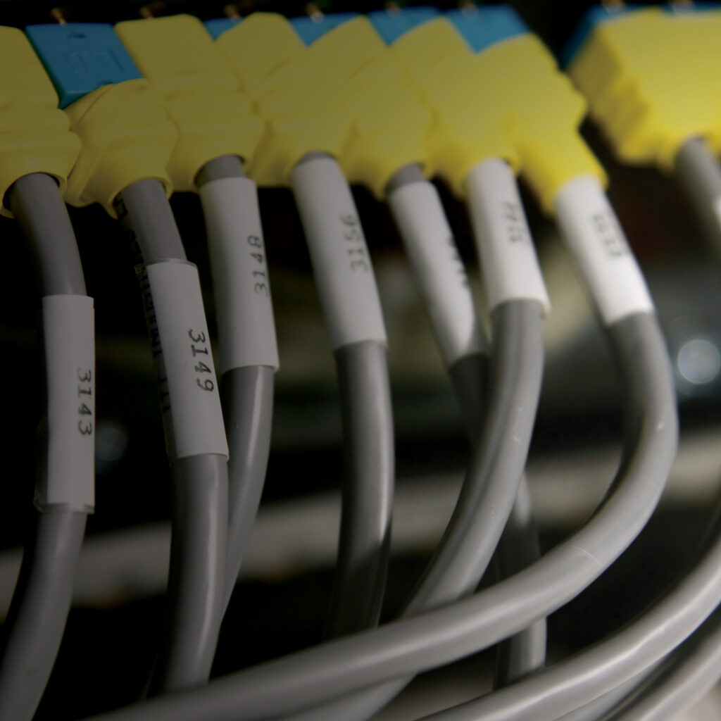 Gray cables with yellow and blue connectors plugged into a panel, each labeled with numbered white tags, against a dark blurred background.