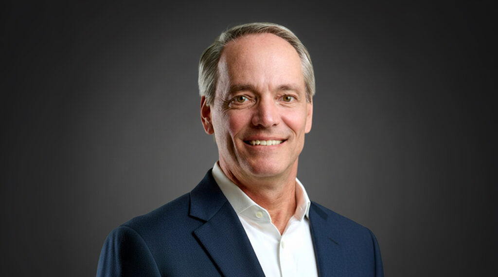 Middle-aged man with short gray hair smiling, wearing a dark blue suit and white shirt against a plain dark gradient background.