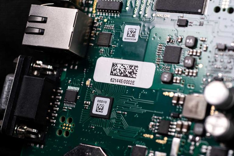 Green circuit board with chips, connectors, ports, QR code labels, and visible circuit pathways.