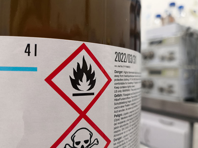 Close-up of chemical label with flammable hazard and partial skull and crossbones; blurred lab equipment in the background.