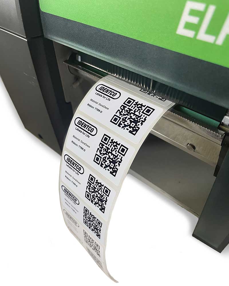 Label printer dispensing a roll of adhesive labels with QR codes, text, and Identco logo, labels partially fed from the machine.