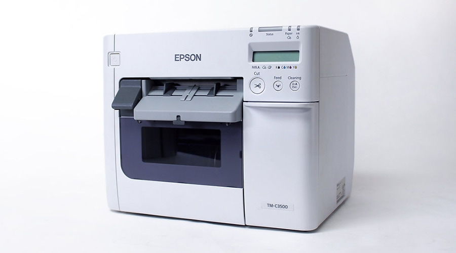 White Epson label printer featuring a digital display, control buttons, and front-loading slot on a plain white background.