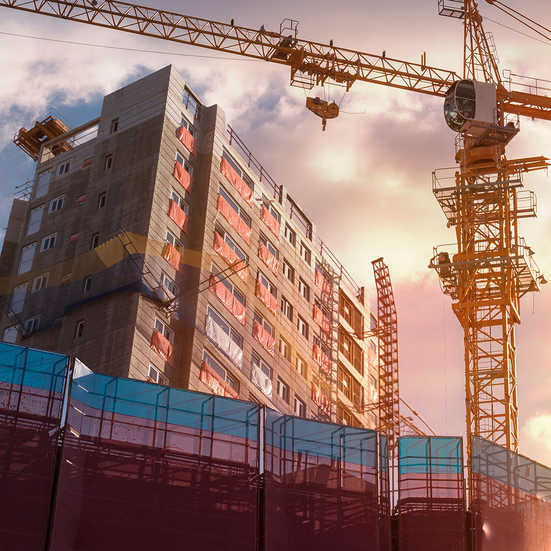 A tall building under construction with cranes nearby and protective netting around the site, illuminated by a warm sunset sky.