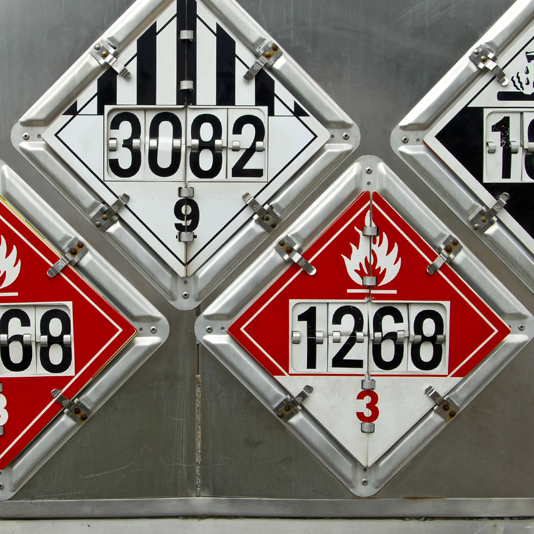 Four diamond-shaped hazardous material placards displayed on a metal surface, showing numbers 3082, 9, 1268, and 3; red placards have a flame symbol, indicating flammable substances.