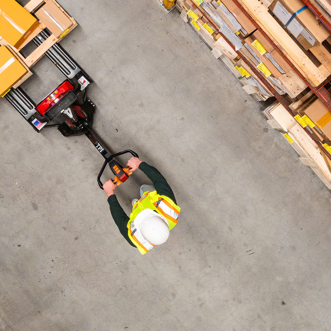 A worker wearing a hard hat and safety vest uses a pallet jack to move boxes in a warehouse, seen from above with shelves of packages to the right.