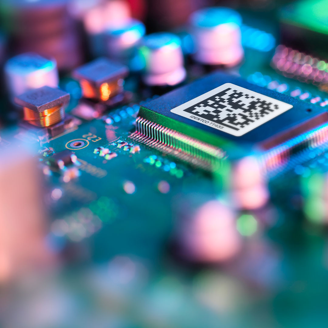 Close-up of an electronic circuit board with a microchip featuring a QR code on its surface, surrounded by colorful blurred components and lights.