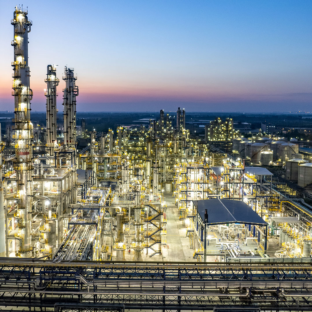 An illuminated oil refinery or petrochemical plant at dusk, with tall distillation towers, industrial structures, and pipes, set against a twilight sky on the horizon.