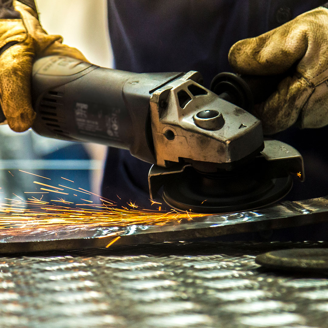 Close-up of a person wearing gloves using an angle grinder on a metal surface, producing bright orange sparks. The focus is on the tool and the metal being worked on.