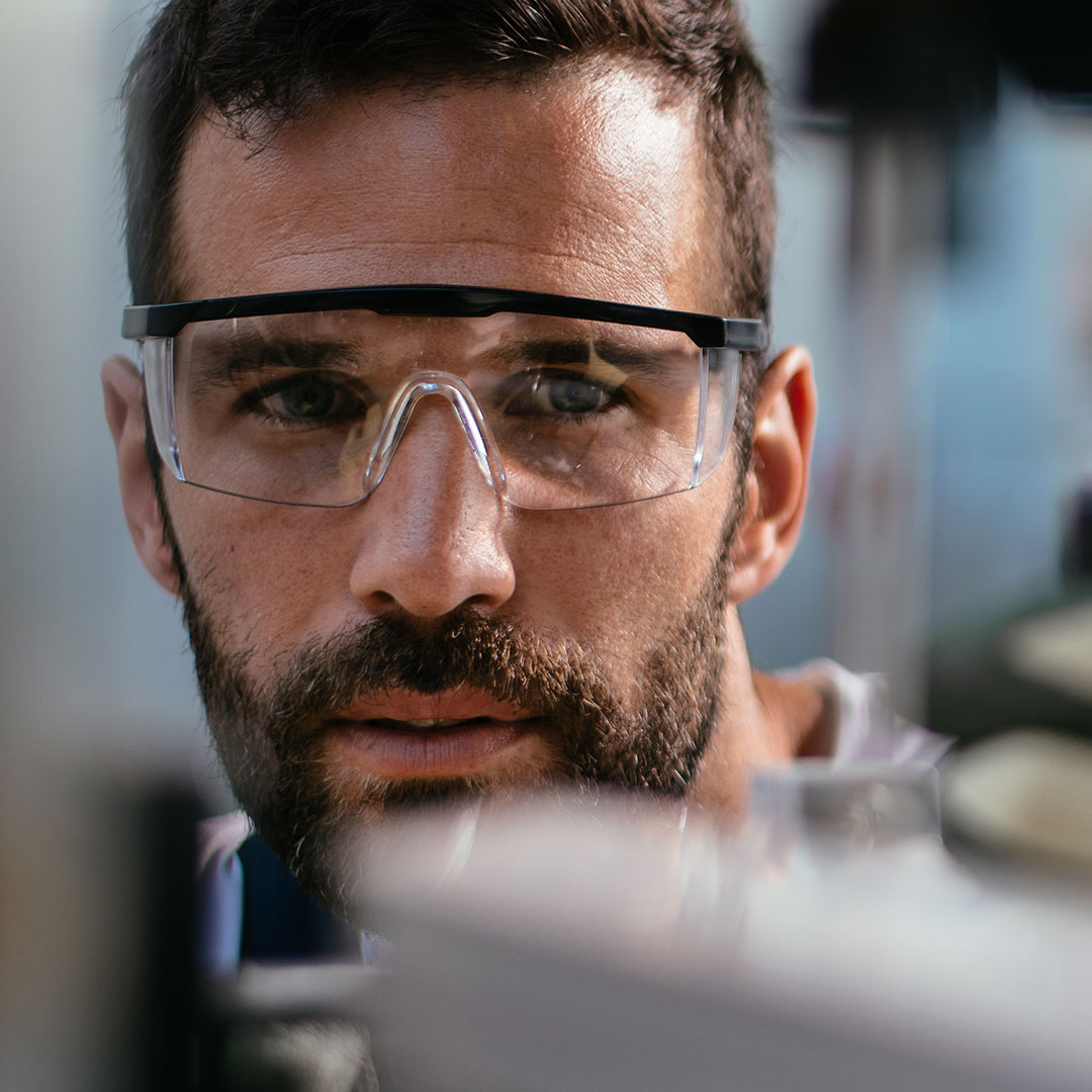A man with a beard wearing clear safety goggles looks intently at something in a lab or workshop environment, with blurred equipment in the foreground.