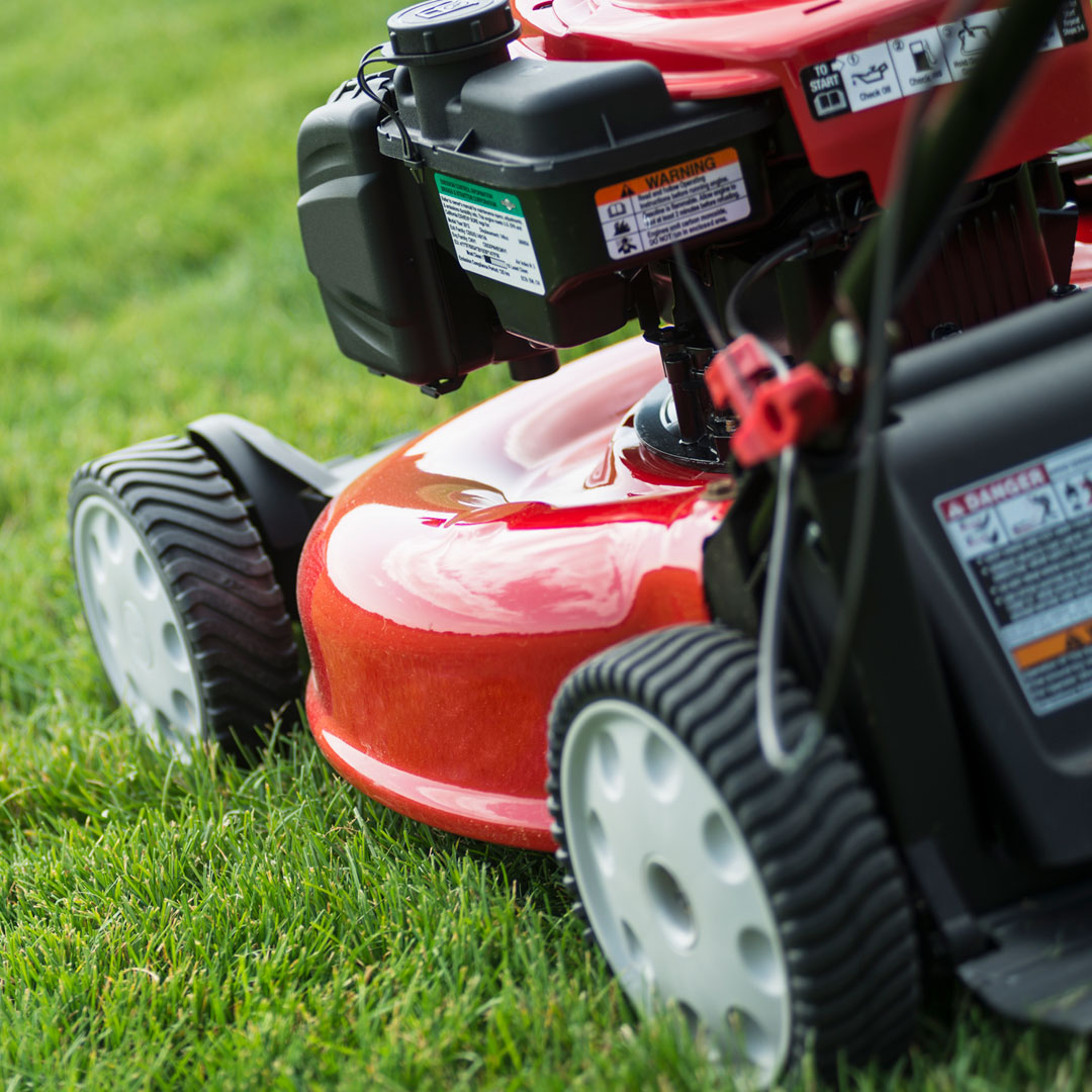 Close-up of a red lawn mower on green grass, showing the wheels, metal deck, and engine with visible warning labels. The image focuses on the front and side of the mower.