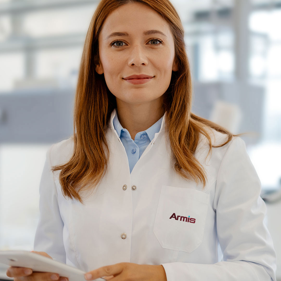 A woman with long brown hair, wearing a white lab coat with the logo Armis on the pocket, stands in a bright laboratory holding a tablet and smiling at the camera.