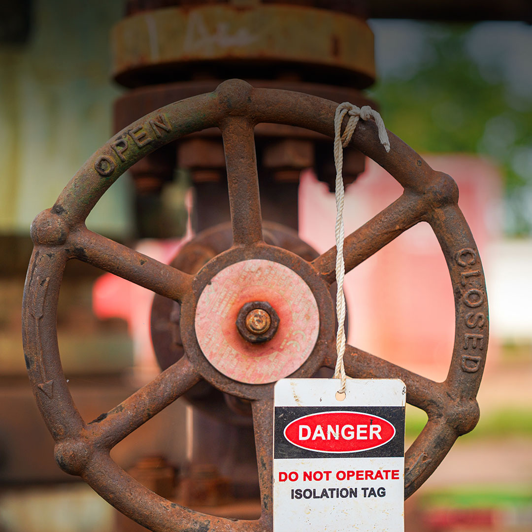 Rusted industrial valve with danger do not operate isolation tag hangs from wheel labeled open and closed, blurred background.