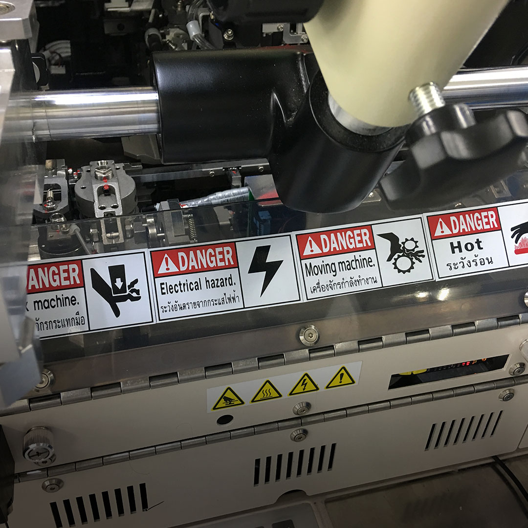 Machine close-up with English and Thai warning labels for electrical hazard, moving parts, hot surfaces, caution symbols, machinery visible.