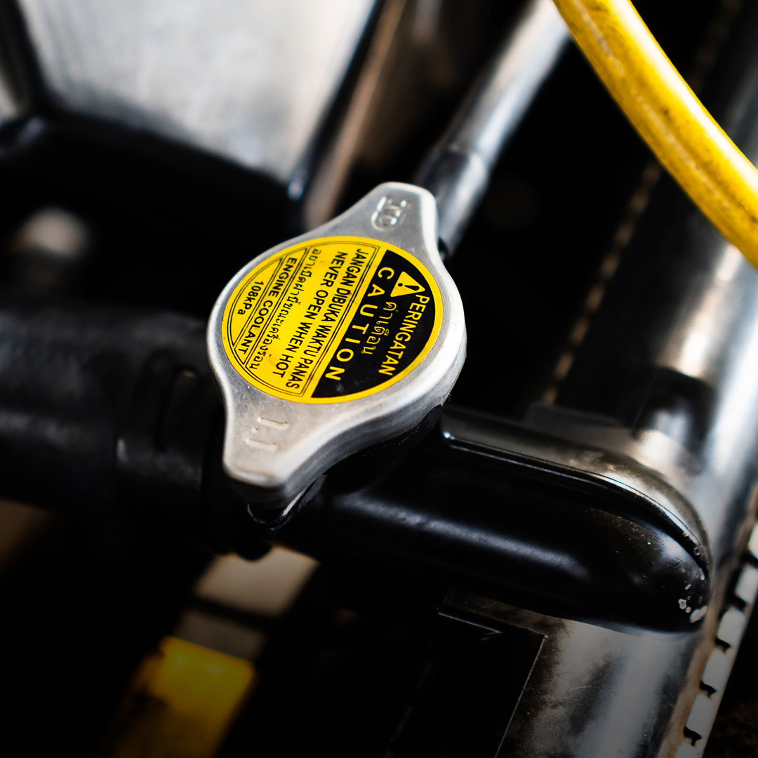 Silver radiator cap with yellow caution label on black car part, yellow hose in background, warning to not open when hot.