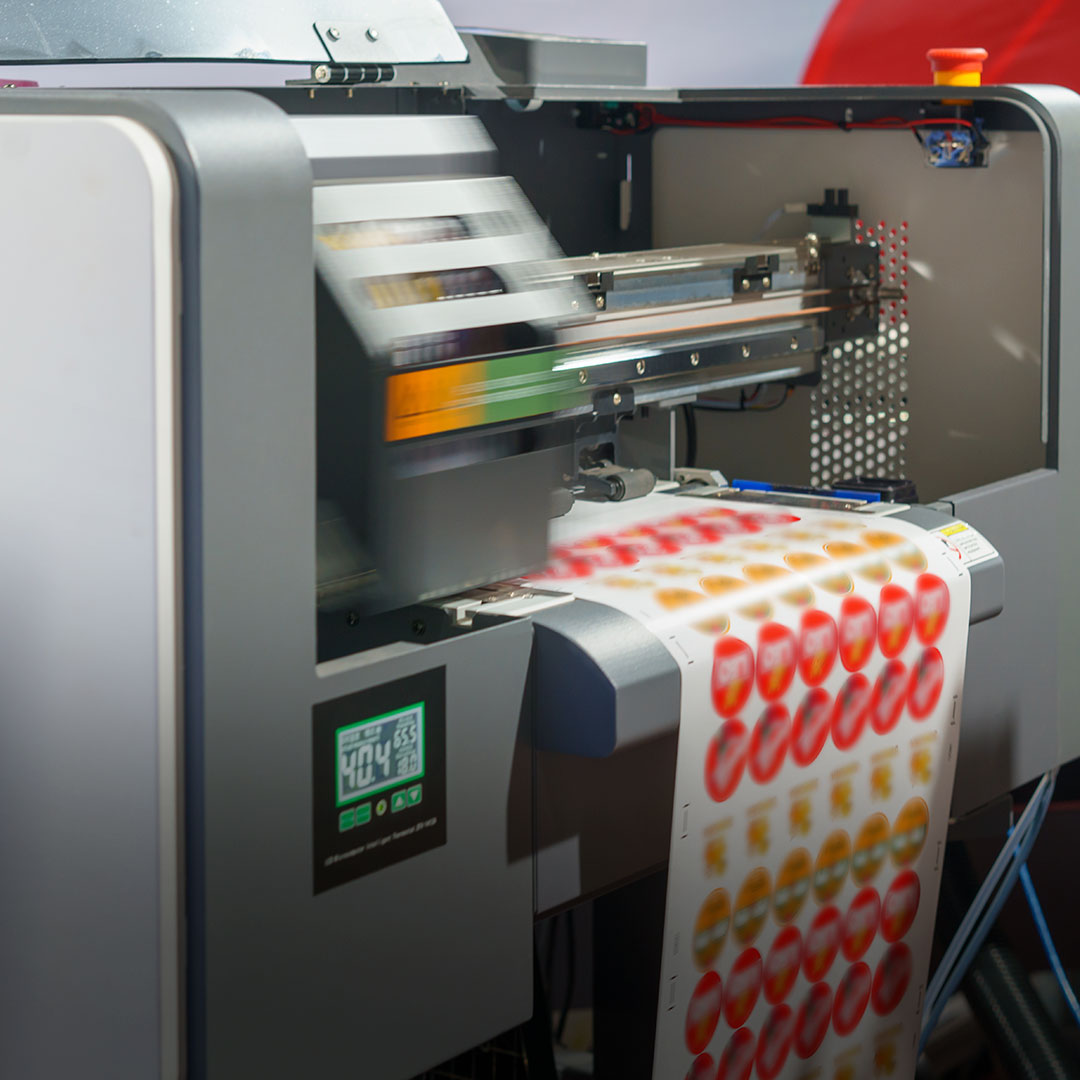 Digital printer outputs colorful sheet labels with red and yellow circles; control panels and indicator lights are on the device.
