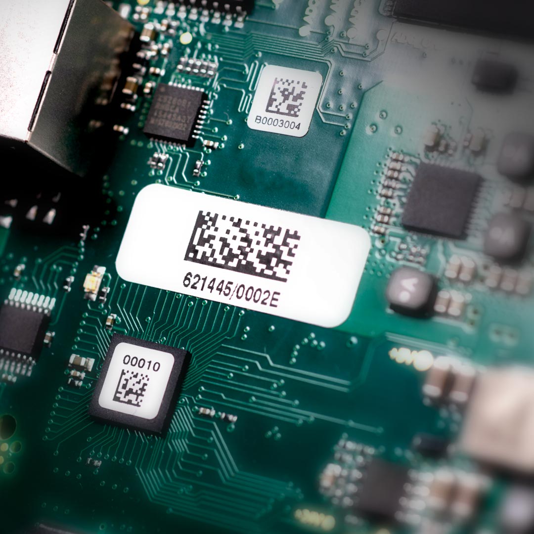 Close-up of a green circuit board with several QR code and barcode labels, electronic components, and microchips visible on the surface.