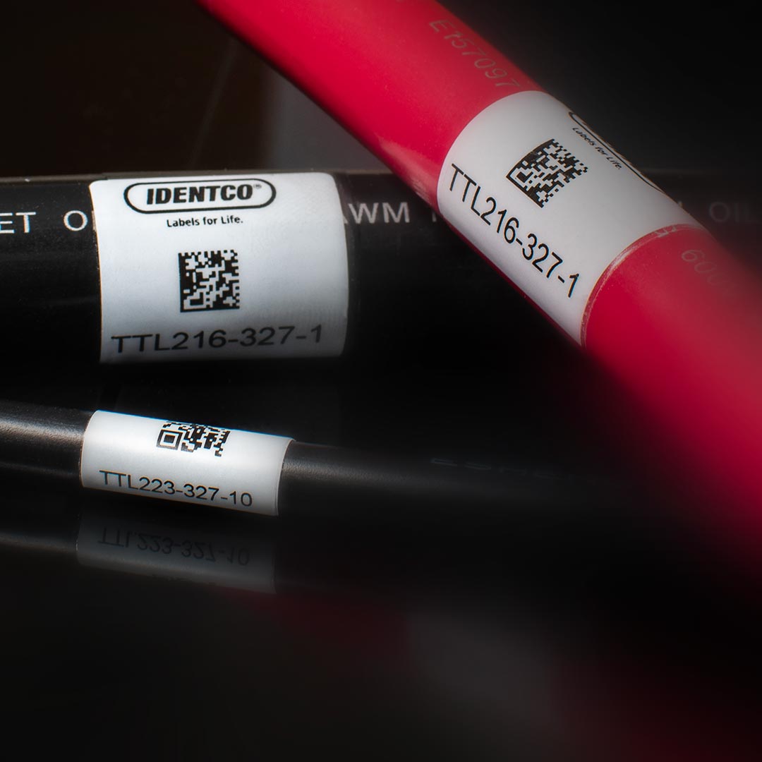 Close-up of three cables with white barcode labels from IDENTCO, each displaying a QR code and serial numbers such as TTL216-327-1 and TTL233-327-10 against a dark background.