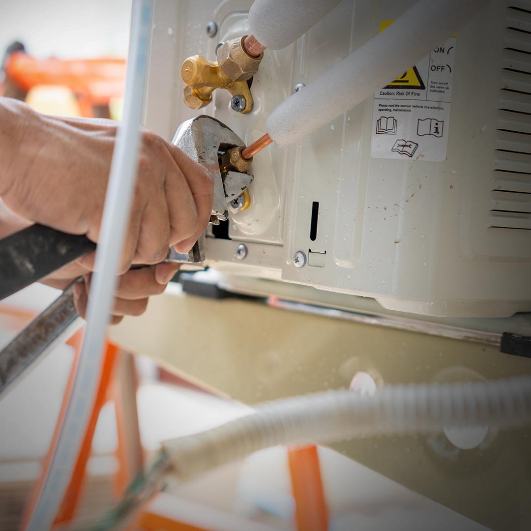 Hands using wrench to tighten pipe on air conditioning unit, surrounded by pipes, tools, and maintenance labels in view.