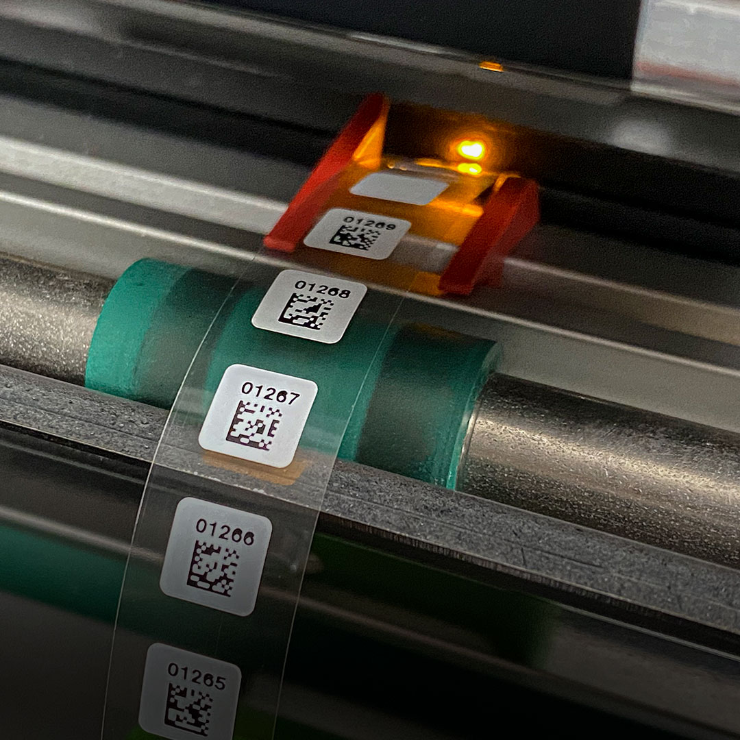 Roll of transparent film with barcode labels, numbers, and QR codes scanned by a machine emitting an orange light.