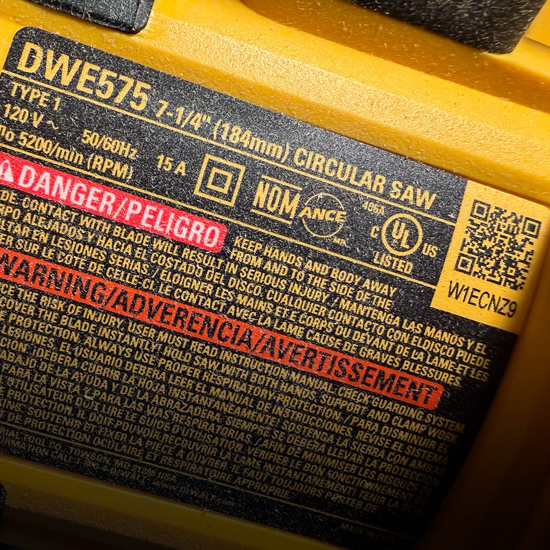 DeWalt DWE575 circular saw label with safety warnings, voltage, amperage, certification icons, and QR code on yellow casing.