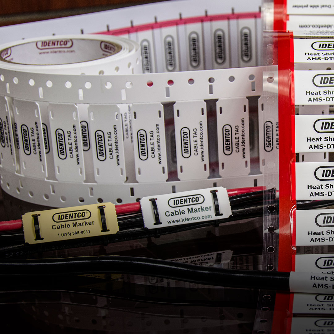 Close-up of IDENTCO cable markers and heat shrink sleeves on rolls and applied to black and red cables with labels and contact info.