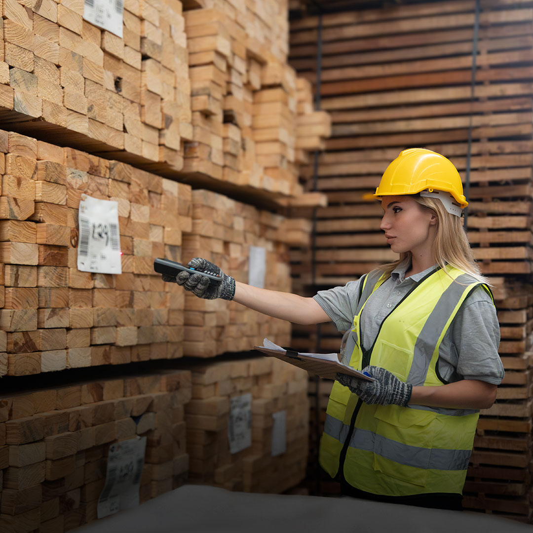 Woman in yellow hard hat and safety vest scans lumber barcodes in warehouse, holding clipboard and wearing work gloves.