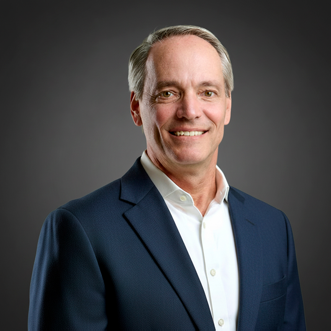 Middle-aged man with short gray hair in a navy suit and white shirt smiles while standing against a dark gray background.