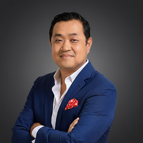 Man in blue suit, white shirt, and red pocket square stands with arms crossed, smiling at camera against dark grey background.