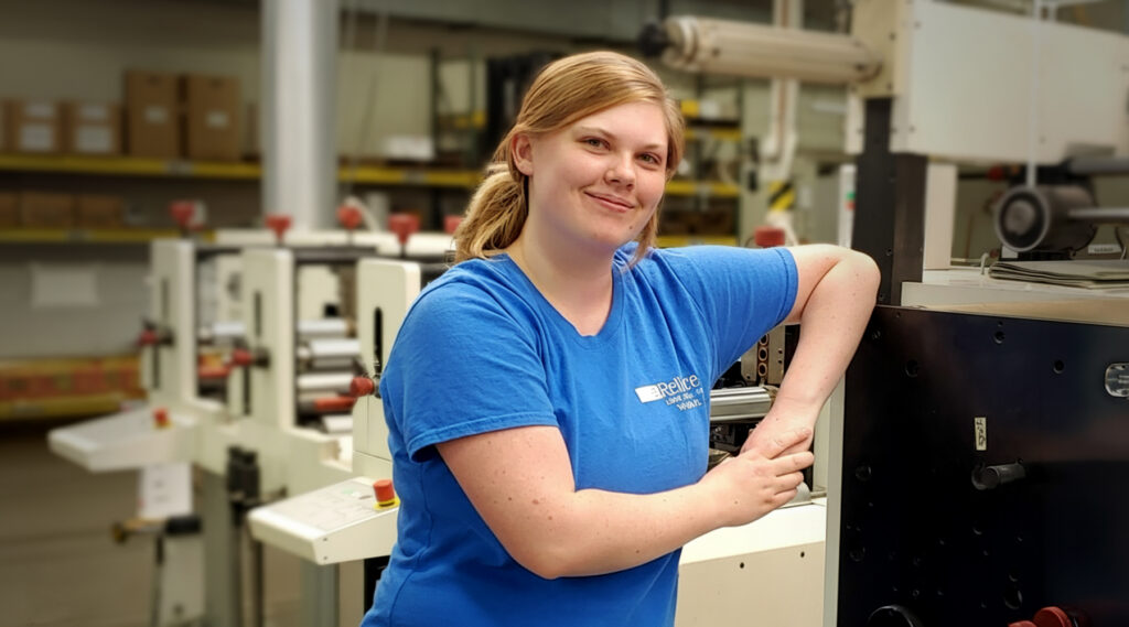 Woman in blue t-shirt smiling and leaning on a black machine in a factory, shelves and equipment visible in the background.