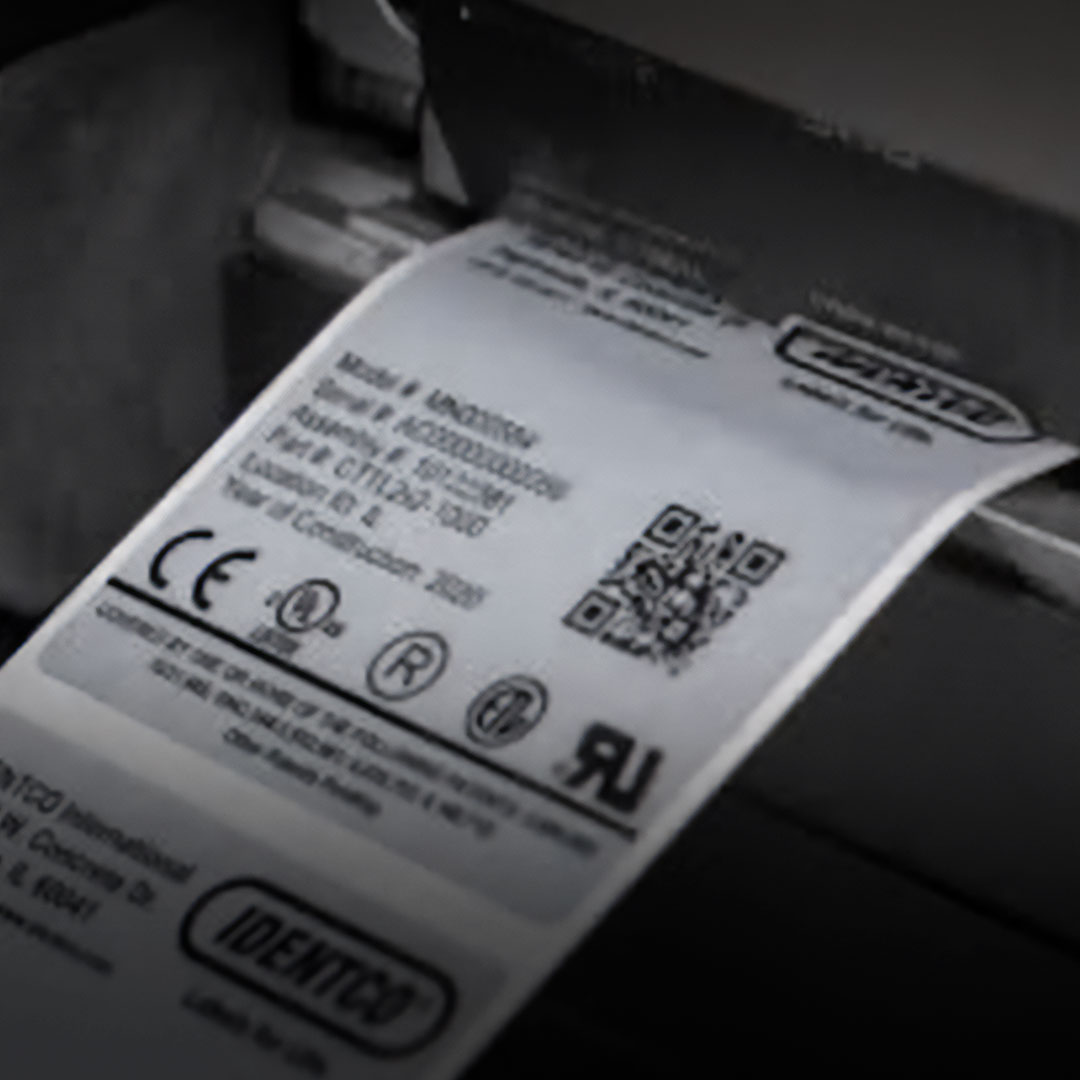 Printer printing a white label with barcodes, QR code, CE and UL logos, certification symbols, and compliance information.