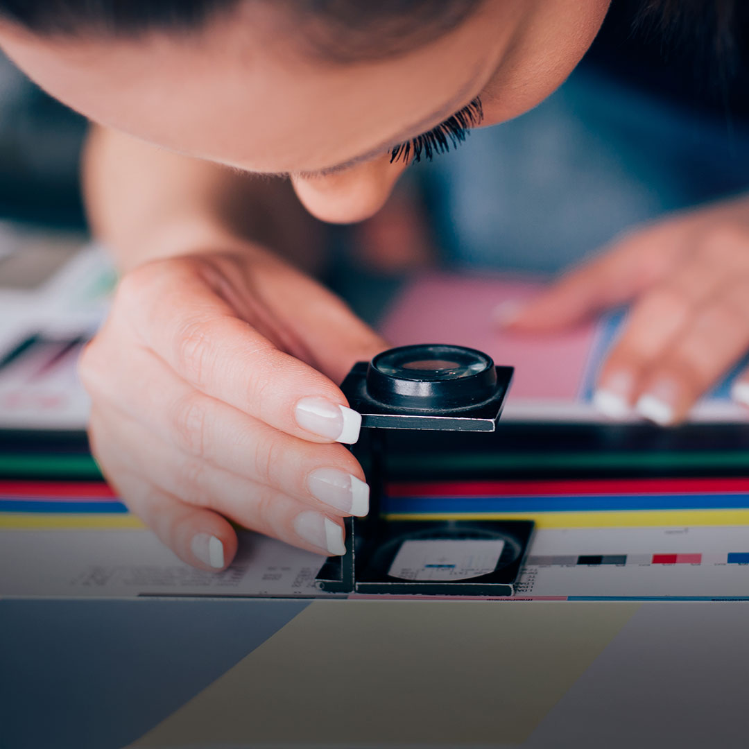 Person uses magnifying glass to inspect color accuracy and print quality on a color sheet, holding it with manicured hand.