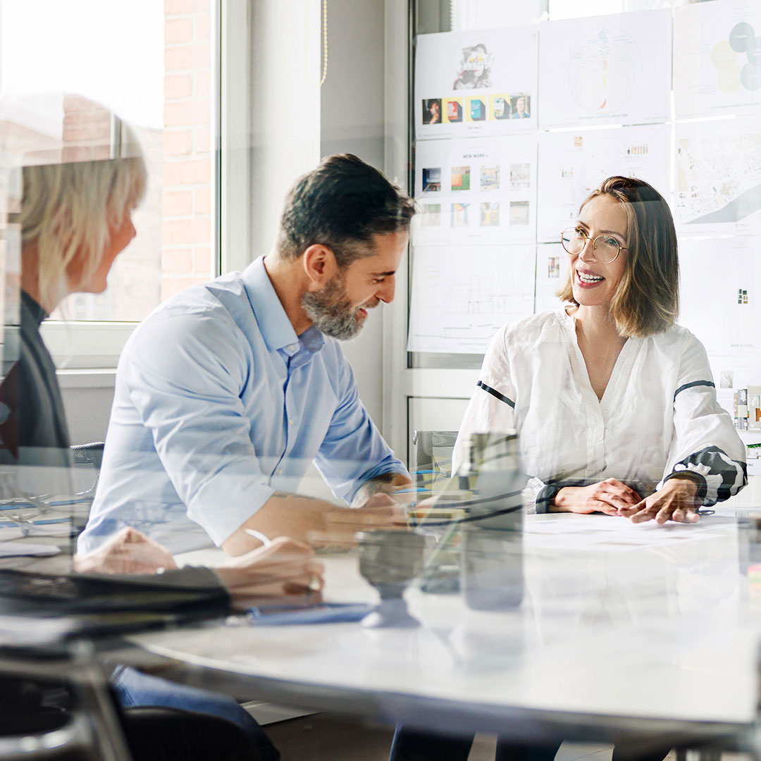 Three colleagues discuss ideas at a table with papers and a laptop in a bright office, design sketches visible on the wall.