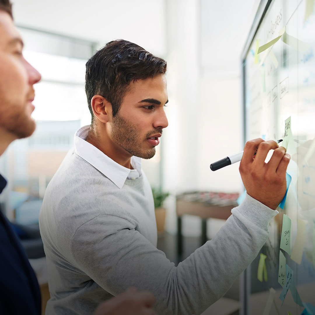 Two men brainstorm in an office; one writes on a glass board covered with sticky notes and diagrams as the other observes.