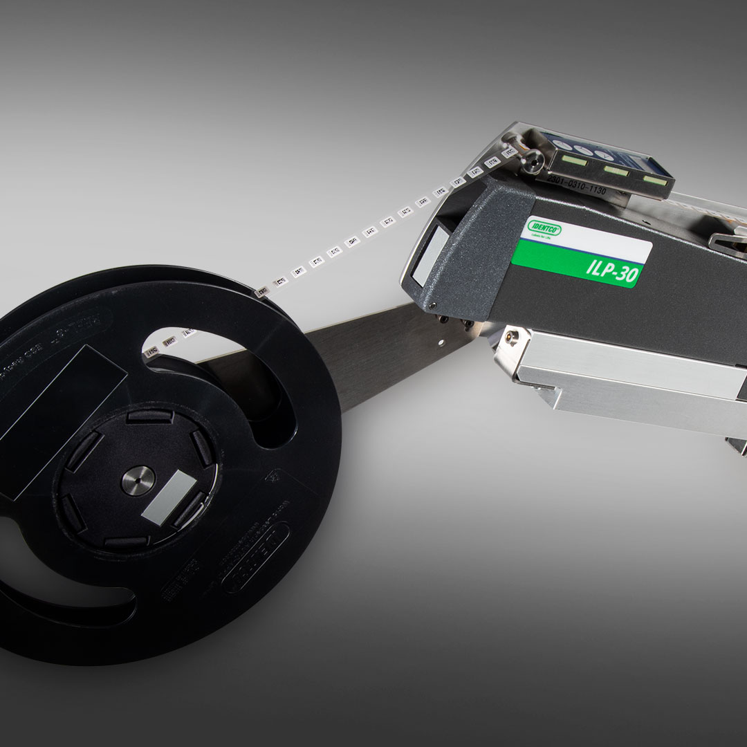Close-up of SMD ILP-30 industrial label printer with black tape spool, set against a gradient gray background.