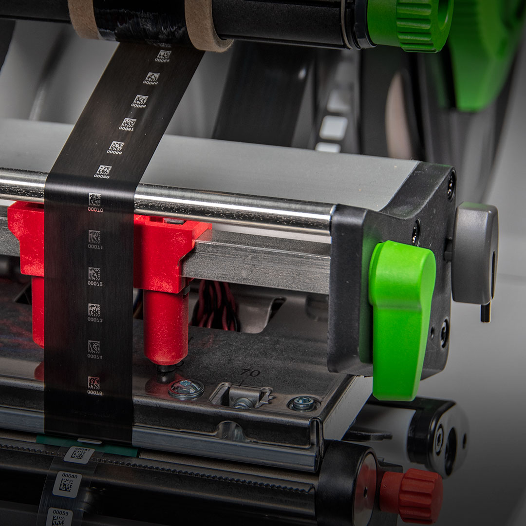 Close-up of a mechanical device with black ribbon icons, metal parts, and red and green levers, likely from a printer or labeler.
