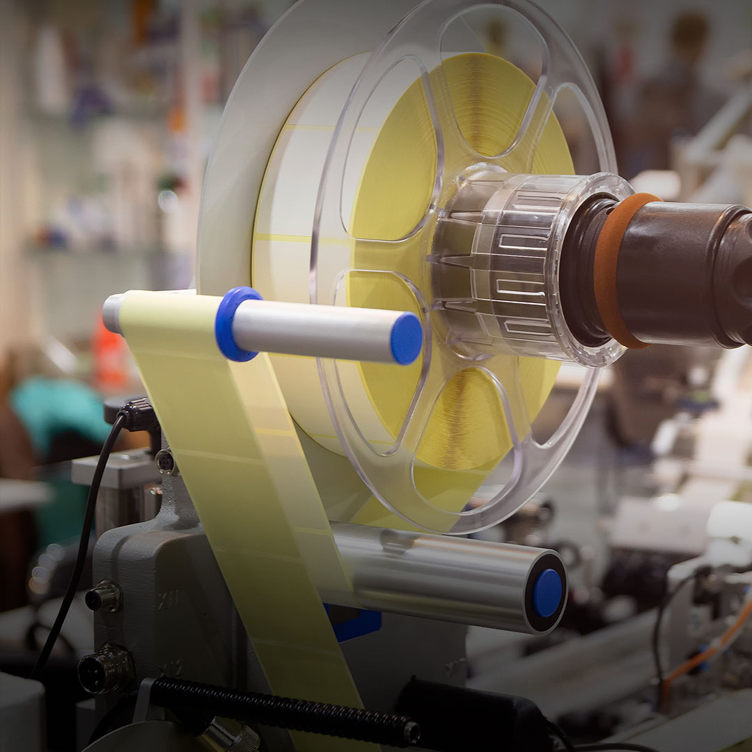 Close-up of a packaging machine unwinding a large yellow adhesive tape spool and applying tape to products in an industrial facility.
