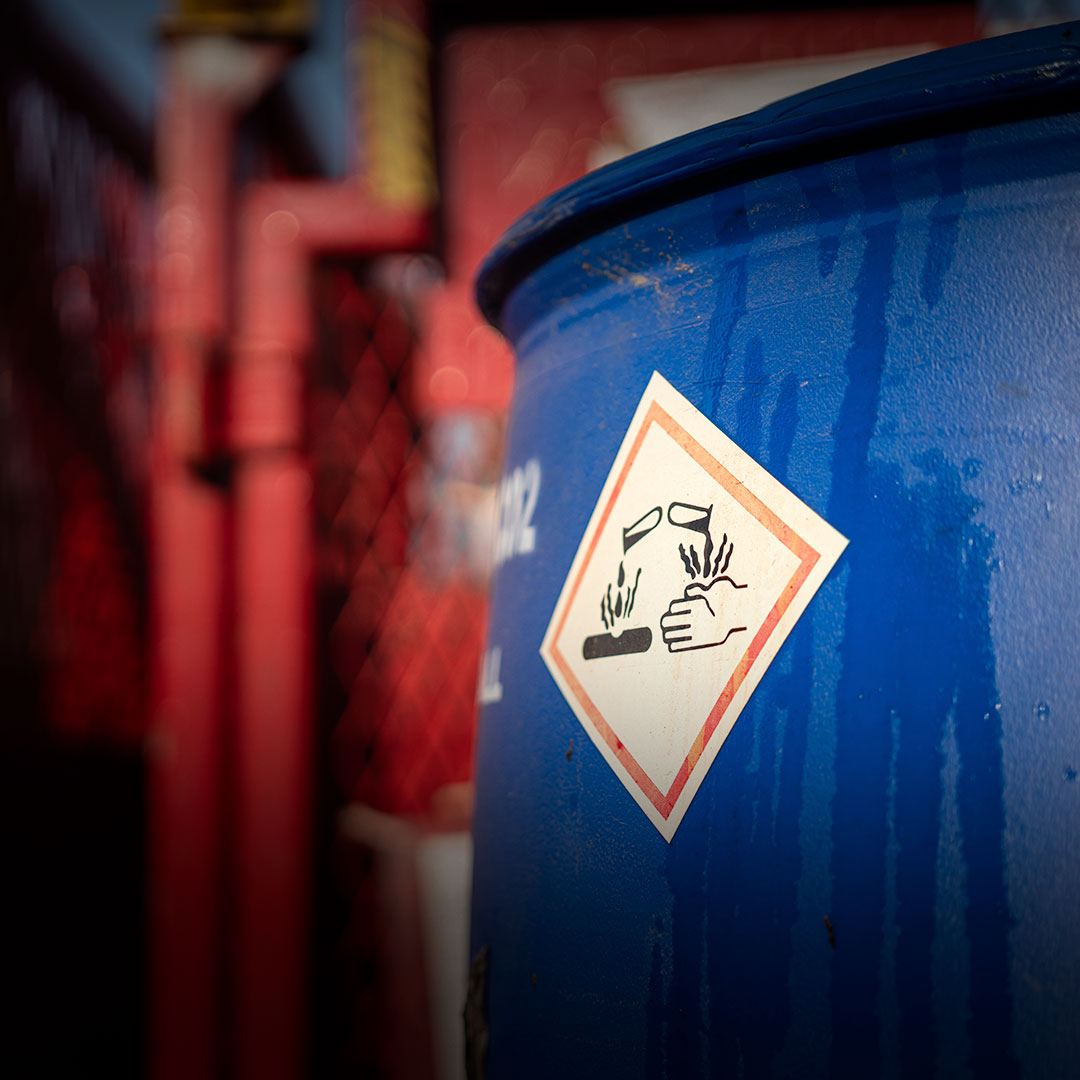 Blue barrel with corrosive warning label, depicting substance damaging hand and metal bar; red pipes and fencing blurred behind.