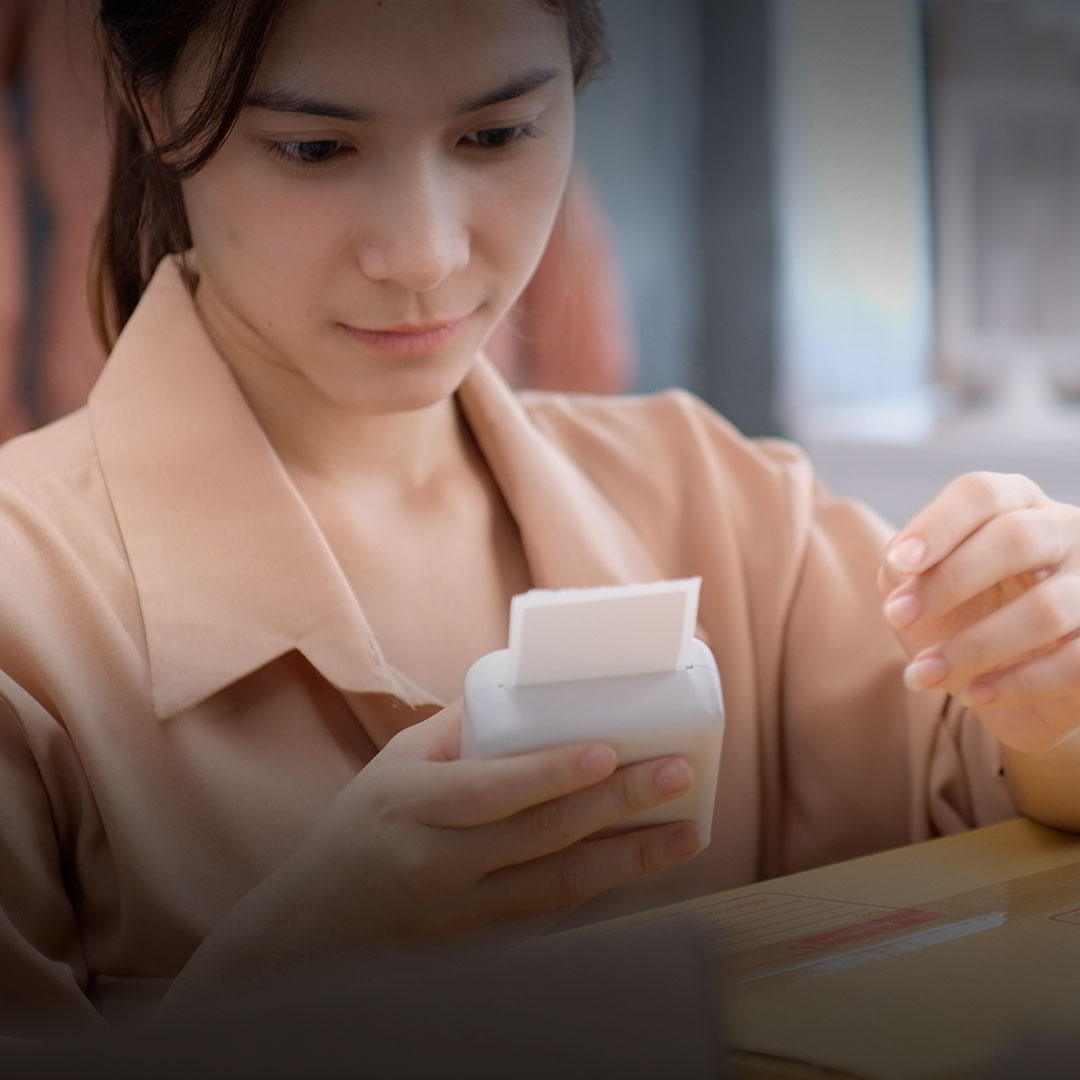 Woman in beige shirt examines small white label maker, holding printed label, seated at desk with cardboard box.