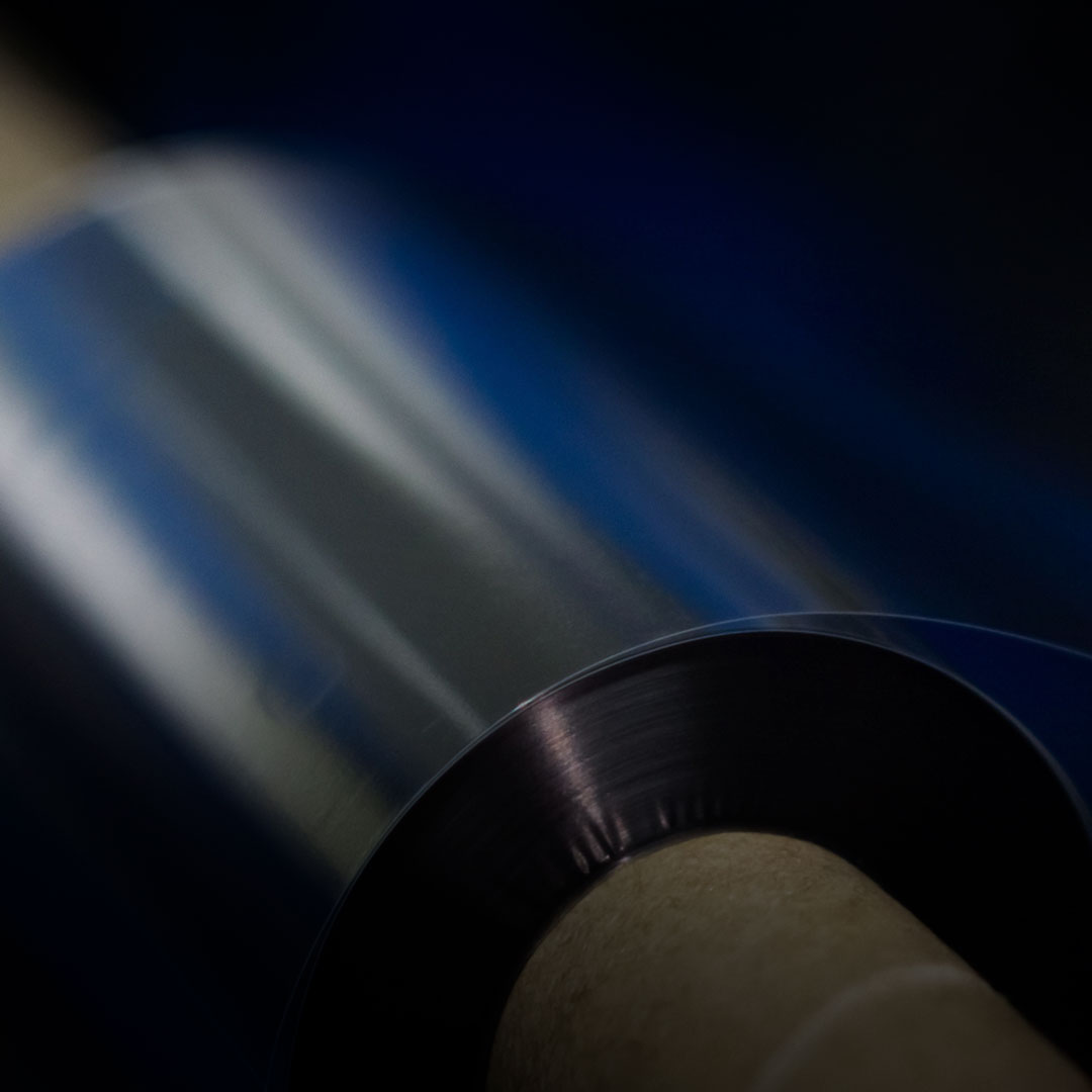 Shiny metallic foil being unrolled from a cylindrical core, close-up with blurred blue and black background, reflective surface.