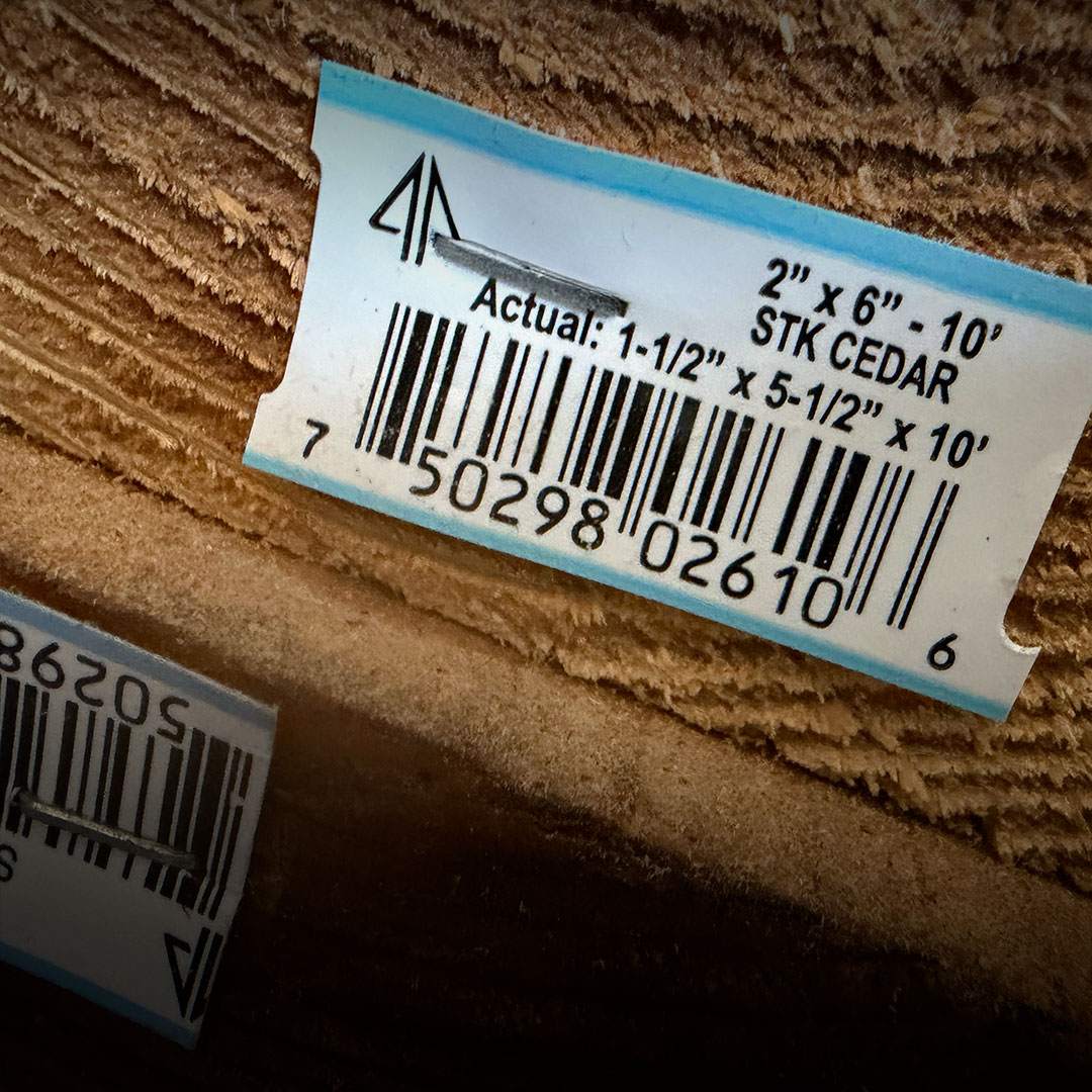 Lumber barcode label for 2x6x10 STK Cedar, actual size 1.5x5.5x10, attached to stacked rough wood boards.