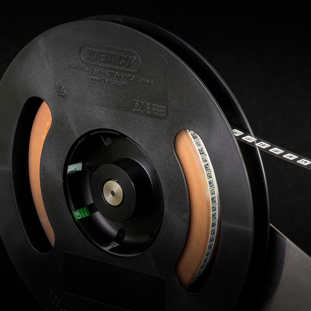 Black plastic reel with spiraled adhesive barcode labels, white with black barcodes and numbers, on a dark background.