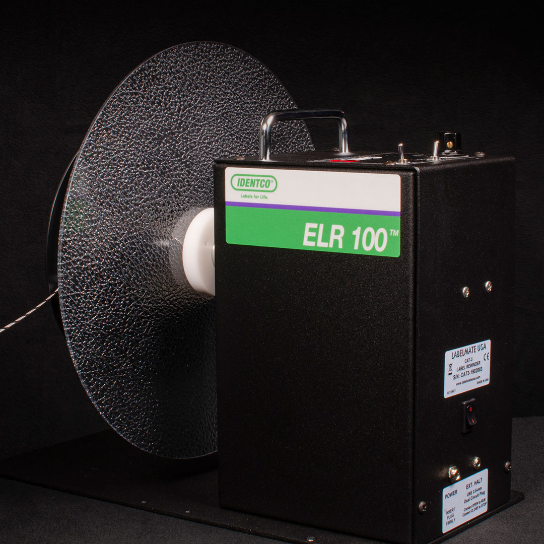 Black and green IDENTCO ELR 100 labeling machine with textured label roll, handle, and labels on surface against dark background.