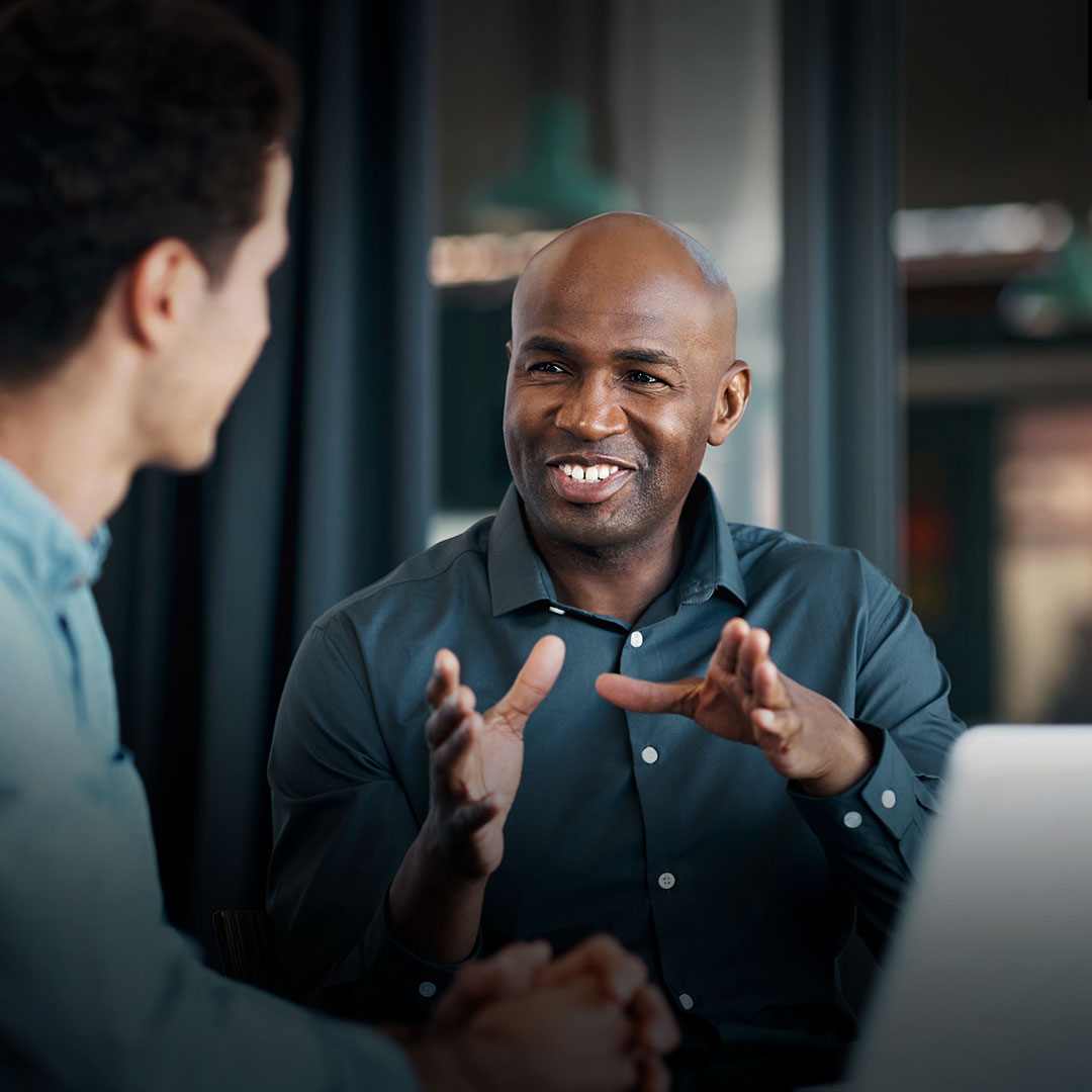 Two men converse at a table, one smiling and gesturing while the other listens attentively; laptop open in the foreground.