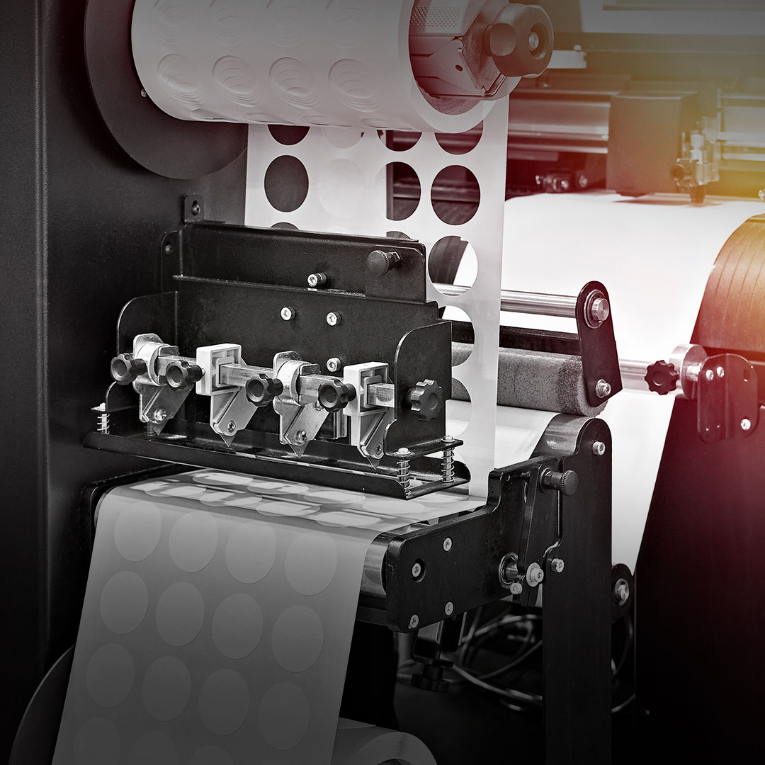 Label printing machine applies adhesive circular stickers to paper roll, with visible mechanical parts and printed labels in factory setting.